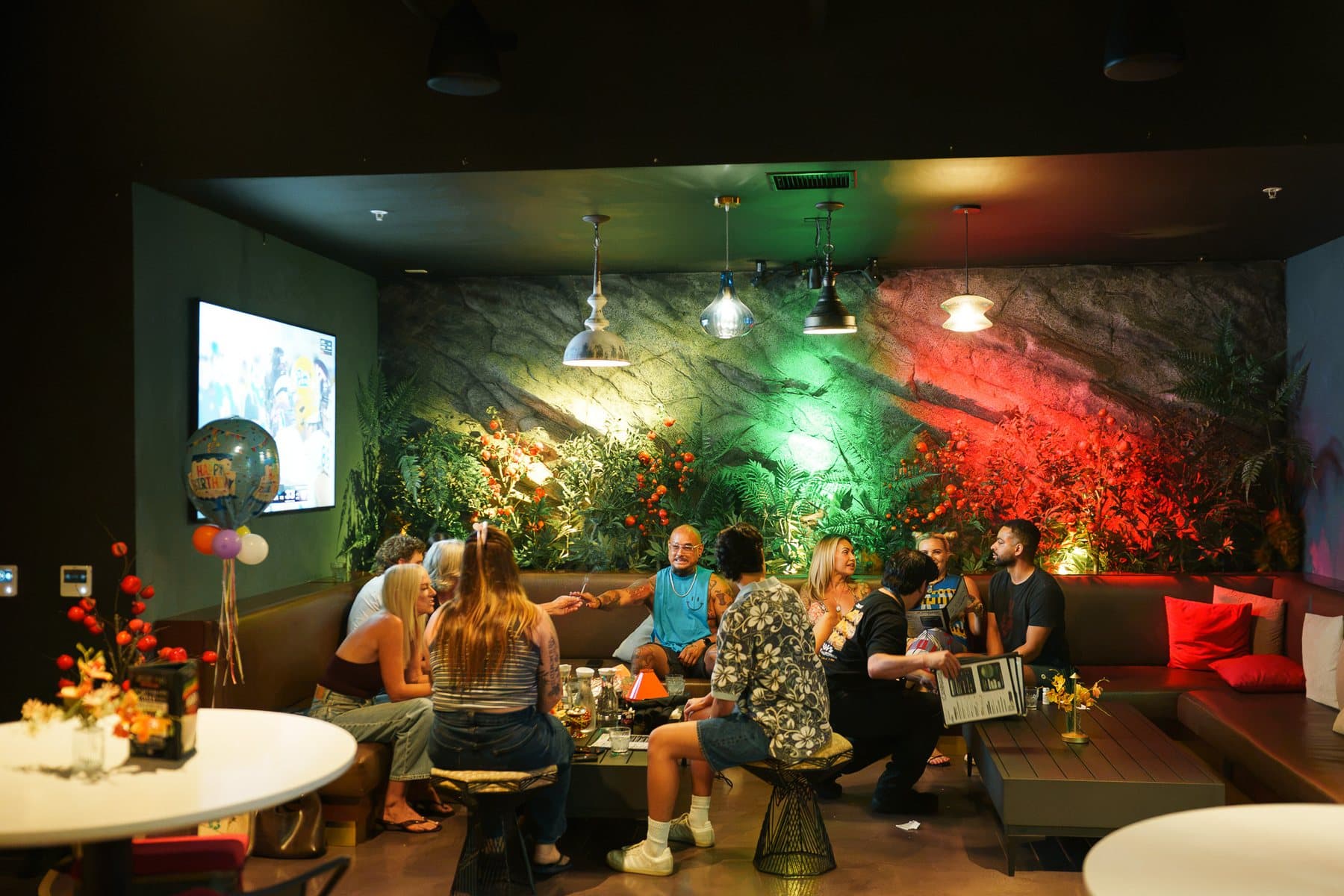 A birthday group on the wraparound banquette in The Den, under pendant lamps and a fern-covered rock wall lit green and red