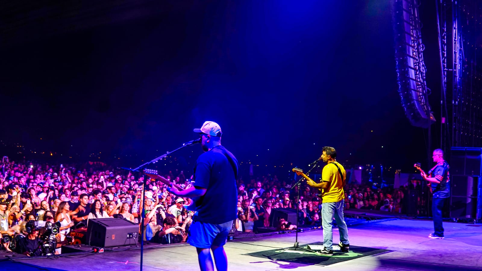 A band plays under blue stage lights to a packed Mission Bayfest crowd, seen from the side of the stage at night