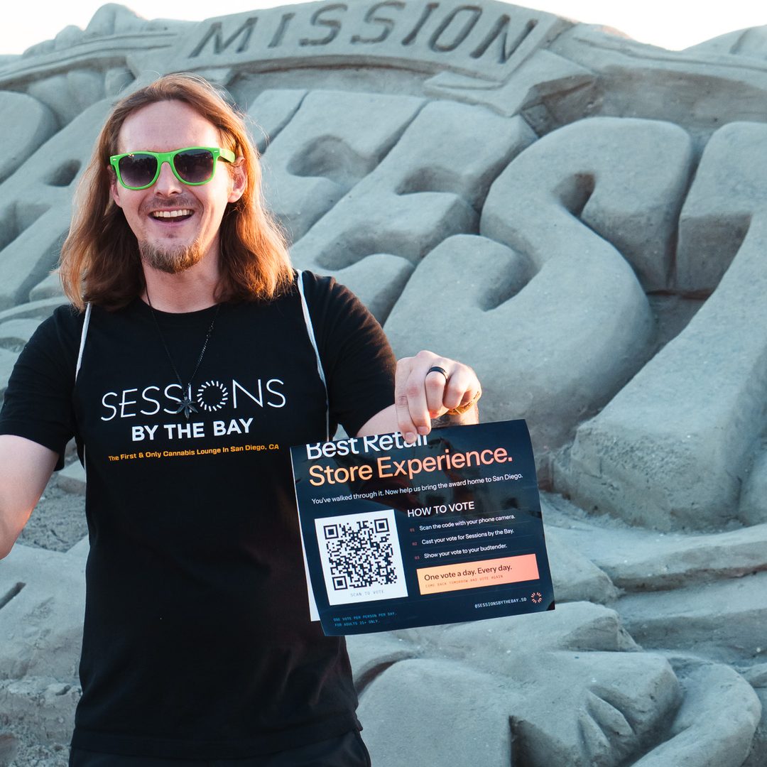 A Sessions team member in green sunglasses holds up an Emjays vote card for Best Retail Store Experience in front of the Mission Bayfest sand sculpture