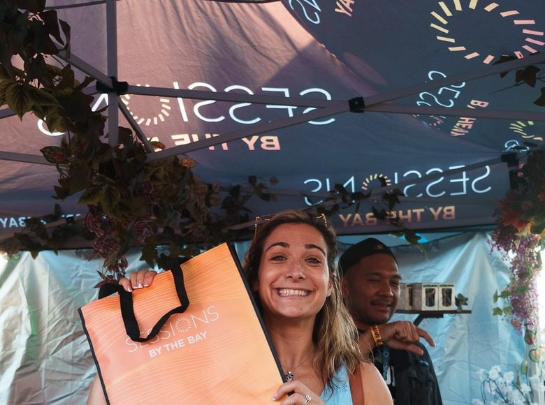 A festival guest smiles under the Sessions canopy, holding up an orange Sessions By The Bay tote bag